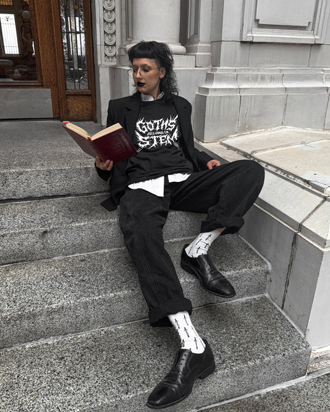 Person sitting on steps reading a book, wearing a black coat and pants with a tshirt that says Goths Belong in STEM in black metal style lettering