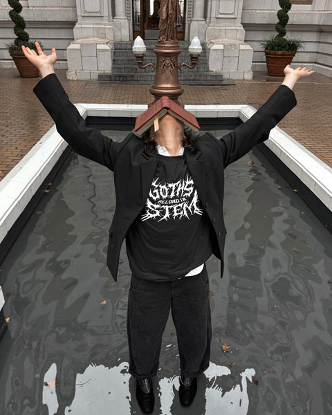 Person standing in foundation with a book resting on their face, both arms up towards the sky wearing a tshirt that says Goths Belong in STEM in black metal style lettering